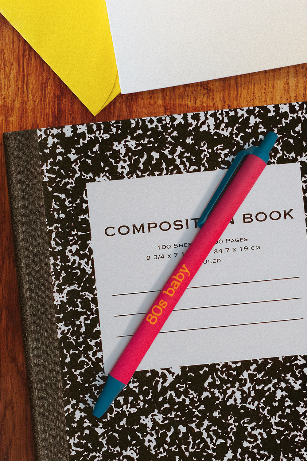 A pink and teal pen with '80s baby' printed on it rests diagonally across a black and white speckled composition notebook on a wooden desk, with a yellow envelope and white paper partially visible.
