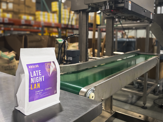 A white coffee bag labeled 'ANALOG LATE NIGHT LAN' with a purple label stands on a stainless steel conveyor belt in a warehouse setting with industrial machinery and shelving in the background.