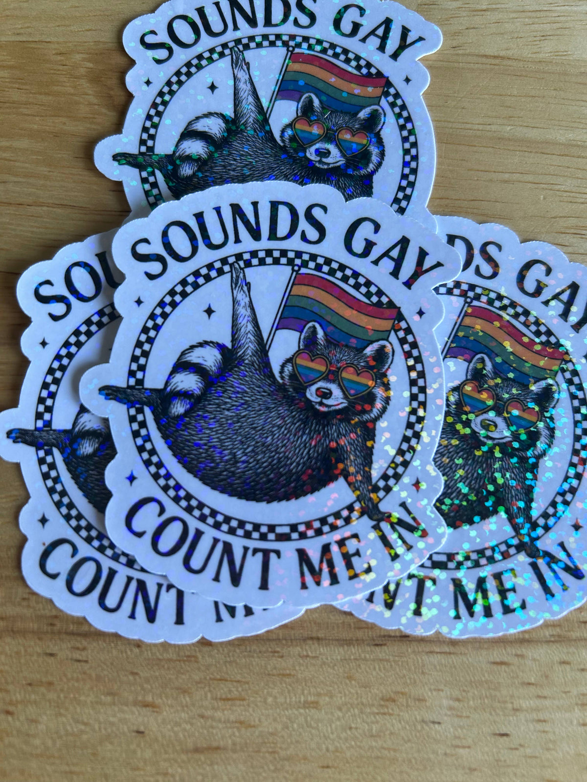 Three die-cut stickers featuring a detailed illustration of a raccoon wearing rainbow sunglasses, with the text 'SOUNDS GAY' and 'COUNT ME IN' on a wooden surface.