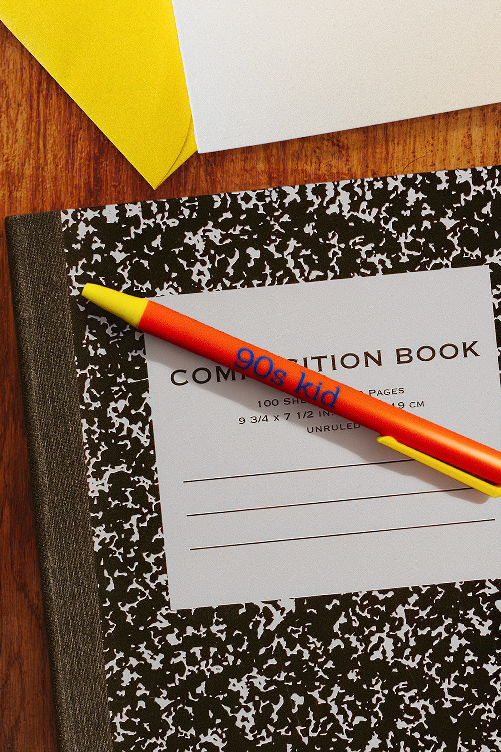 A classic black and white marbled composition notebook on a wooden desk, with a bright orange and yellow pen reading 90s kid resting diagonally across the cover. A white sheet of paper with printed writing is visible underneath, and other desk items are partially cropped at the top.