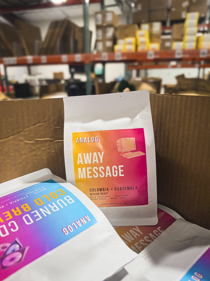 A white coffee pouch labeled 'AWAY MESSAGE' with a pink-to-orange gradient label, sitting inside a cardboard box with other branded pouches visible in a warehouse setting.
