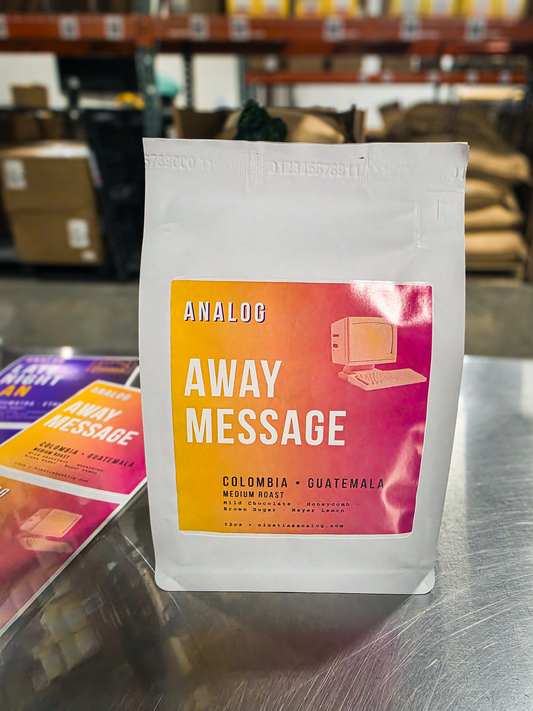 A white stand-up coffee bag labeled Analog Away Message, placed on a metal counter in a warehouse or store setting with shelves and bags of coffee in the blurred background.