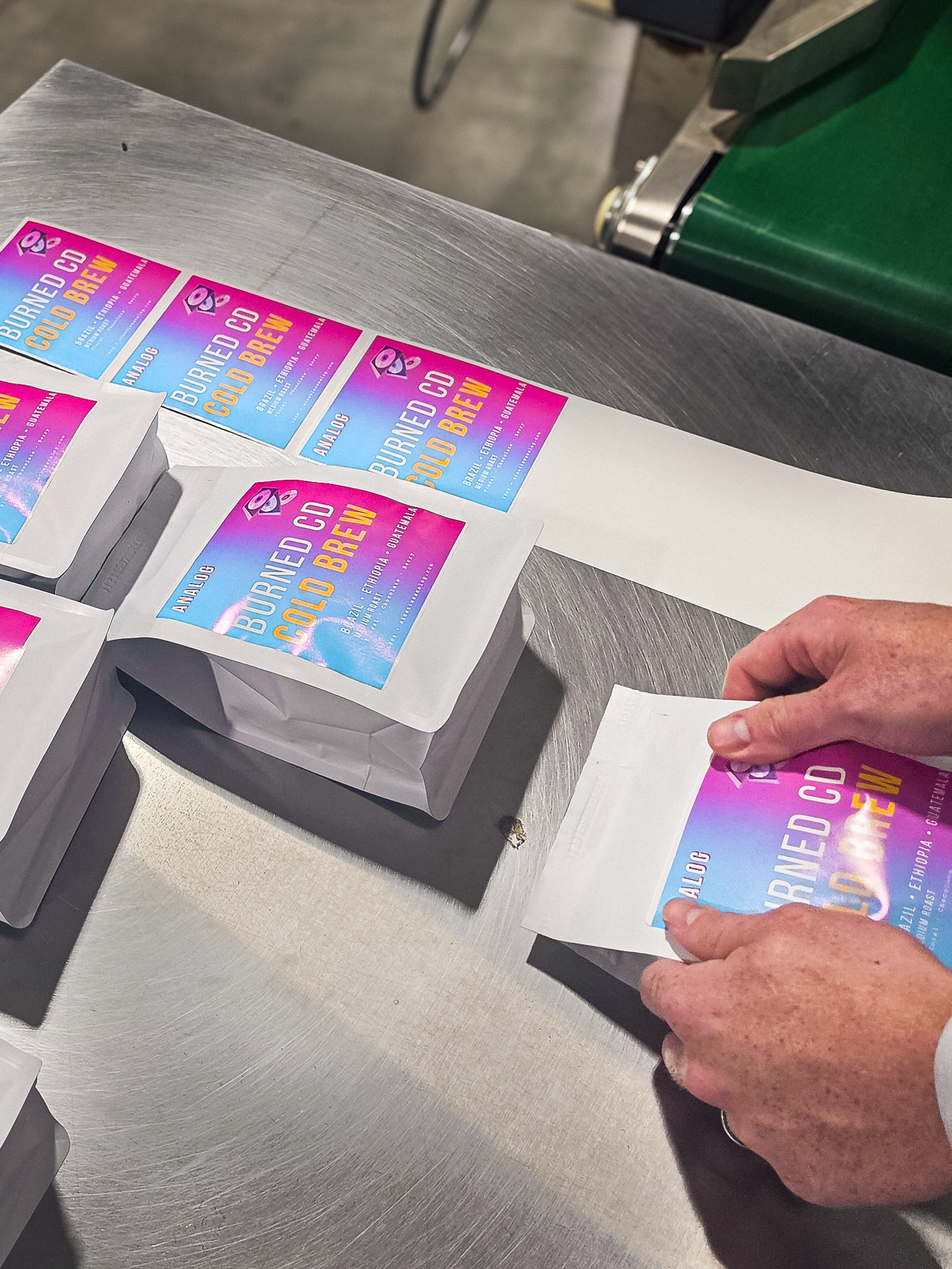 A close-up of a person's hands placing a sticker onto a small white coffee pouch with a colorful blue-to-pink gradient label onto a stainless steel conveyor belt. The label reads 'BURNED CD COLD BREW.'