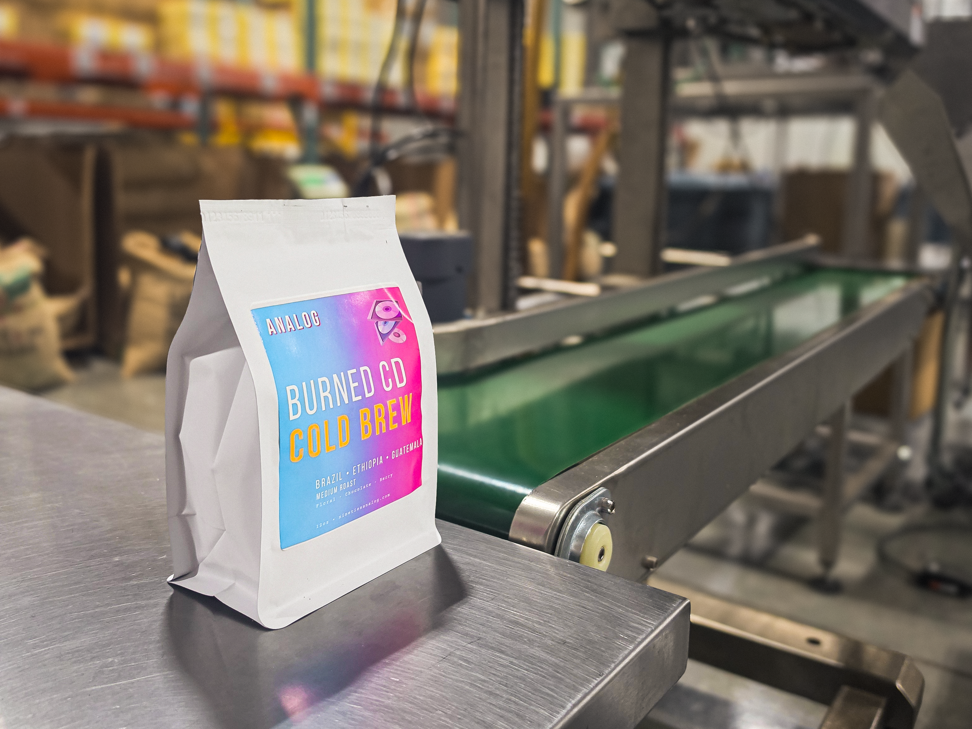 A white stand-up coffee bag labeled 'ANALOG BURNED CD COLD BREW' with a colorful gradient label, standing on a stainless steel conveyor belt in a warehouse setting.