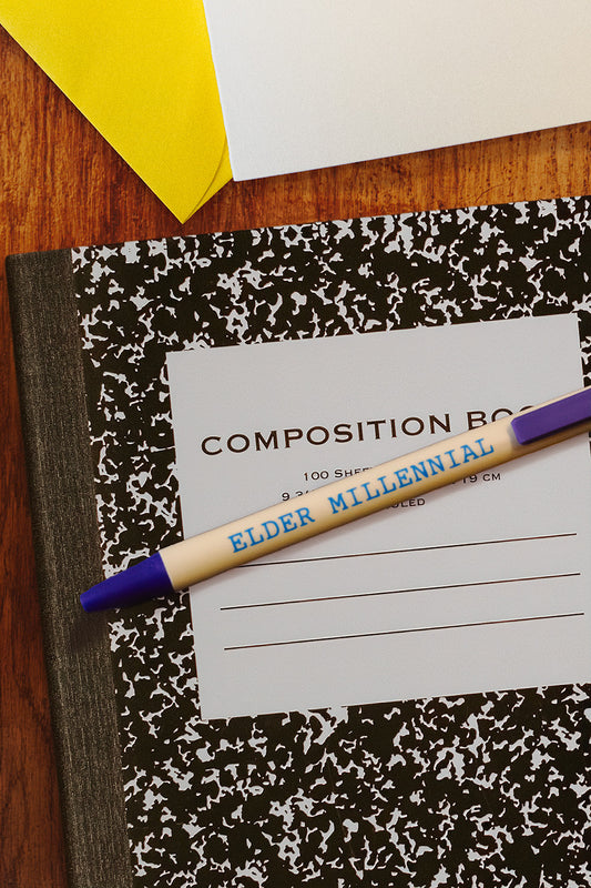 A customized nostalgic ANALOG pen with blue lettering reading 'ELDER MILLENNIAL' and purple accents lies across a classic black-and-white marbled composition notebook on a wooden desk.