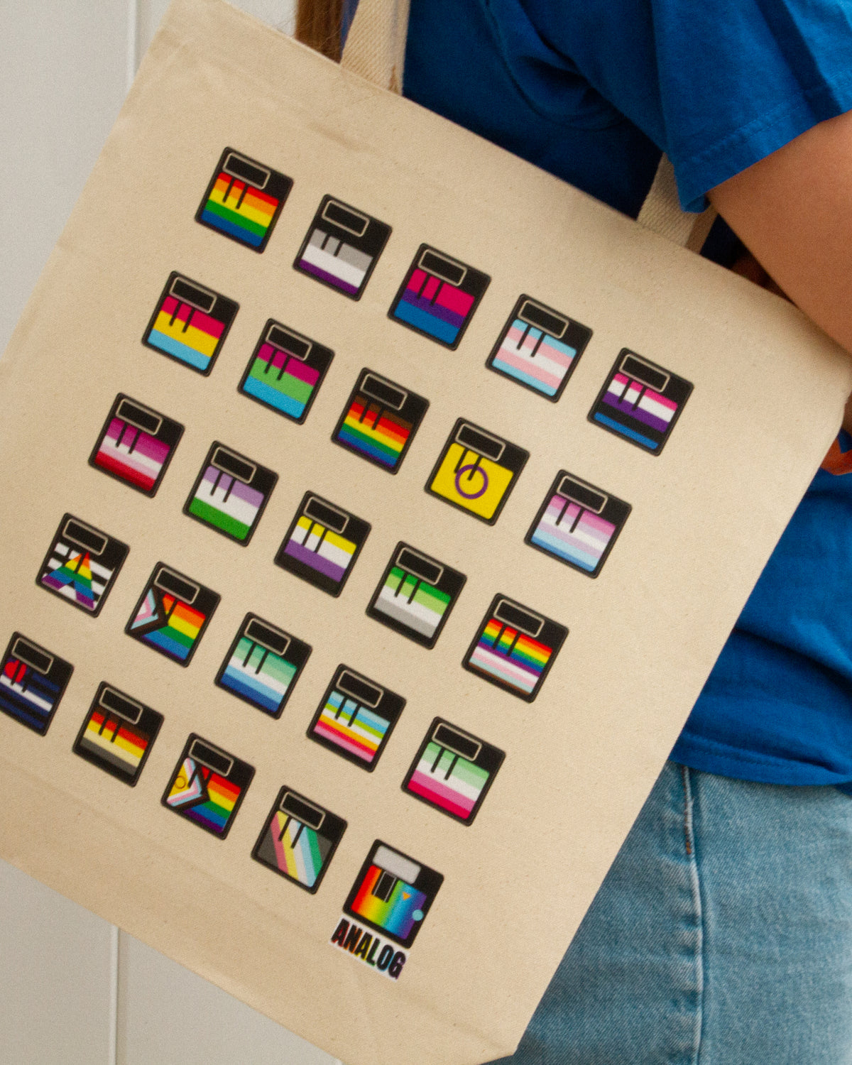 A beige canvas tote bag printed with multiple pride flag floppy disk icons and the word 'ANALOG' at the bottom, carried by a person wearing a blue shirt and jeans.