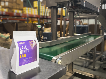 A white coffee bag labeled 'ANALOG LATE NIGHT LAN' with a purple label stands on a stainless steel conveyor belt in a warehouse setting with industrial machinery and shelving in the background.