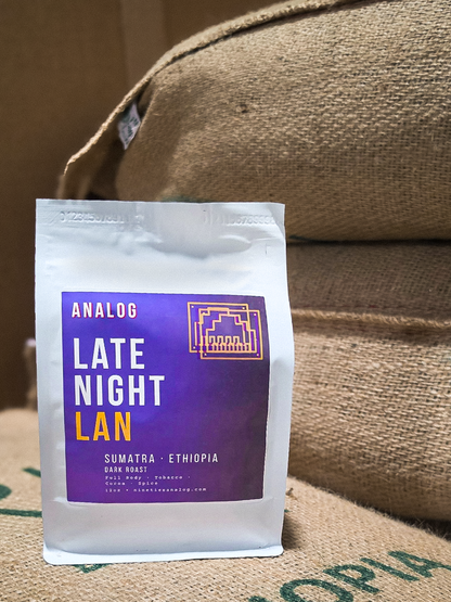 A white bag of Analog Coffee Roasters 'Late Night Lan' Sumatra Ethiopia dark roast coffee, featuring a purple label with white and orange text, set against a background of brown burlap sacks.